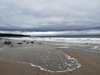 A serene seascape unfolds on a sandy beach, the sky covered with soft, gray clouds that give the scene a calm, overcast atmosphere.