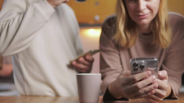 Digital disconnection defining modern couple silently sitting in kitchen, absorbed in smartphones with untouched coffee mugs nearby