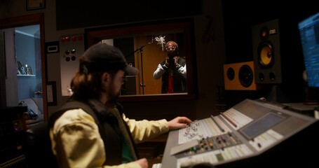 The rapper is recording song in the studio. The sound engineer controls the recording process sitting in the control room