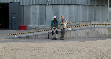 Two workers in uniform are having lunch sitting outside near the factory. They're talking.