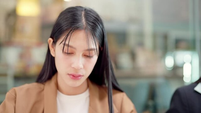 A woman with long hair is sitting at a table with her head down. She is wearing a brown jacket and a white shirt