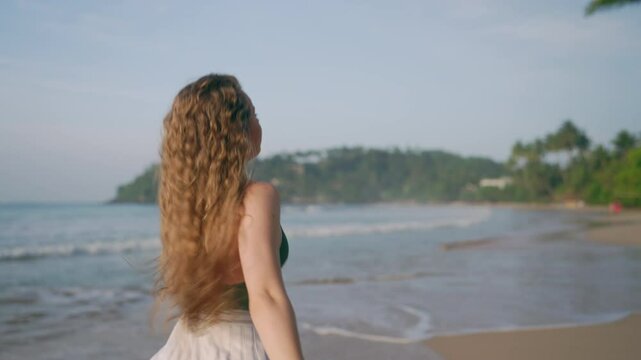 Woman in white dress frolics on tropical beach, lush curls bounce energetically. Solo traveler enjoys seaside, vitality of sun-kissed hair exudes freedom. Hat in hand, back to camera, pure relaxation.