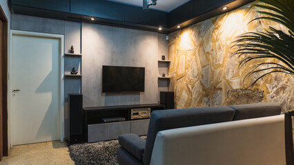 A modern living room with a gray accent wall and a large TV. The room features a comfortable couch, a unique textured wall, and a glimpse of a plant. The space is stylish and well-lit.

