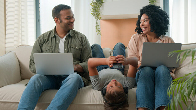 Addicted family use gadgets devices little kid child son boy lying upside down on sofa play video game on mobile phone smiling African American parents working laptops different gen using modern tech - Powered by Adobe