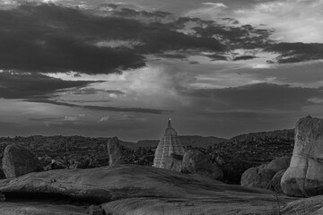 The ancient stone temples of Hampi stand in serene grandeur against a dramatic sunset, reflecting...