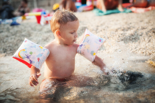 A young child sits in shallow water at the beach, wearing colorful arm floaties. The child splashes water joyfully, surrounded by bright beach toys and cheerful summer crowds.
