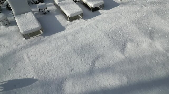 aerial view of snow covered lounge chairs next to closed shuttered swimming pool in winter (back yard patio loungers outdoor furniture) spa bizzard snowy snowfall frozen home countryside drone footage
