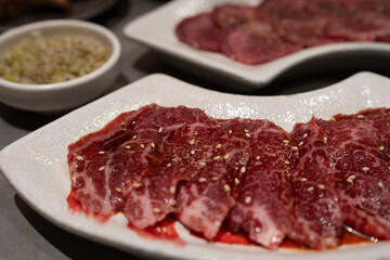 A plate of marbled raw beef slices drizzled with sauce and sprinkled with sesame seeds, ready for grilling. A side of chopped scallions is visible in the background.