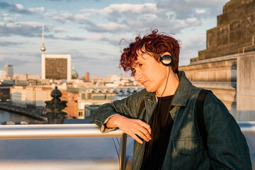 Tourist enjoying berlin cityscape at sunset wearing headphones and leaning on a railing