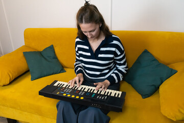Young woman is sitting on a comfortable yellow sofa, playing an electronic keyboard, engrossed in...