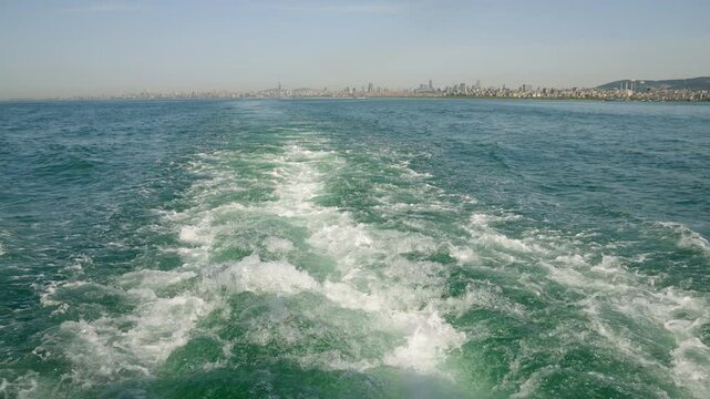 Ferry trail in sea with view of Istanbul skyline. Water waves, urban cityscape in background, sunny day. Sail, travel, explore Turkish culture from boat.