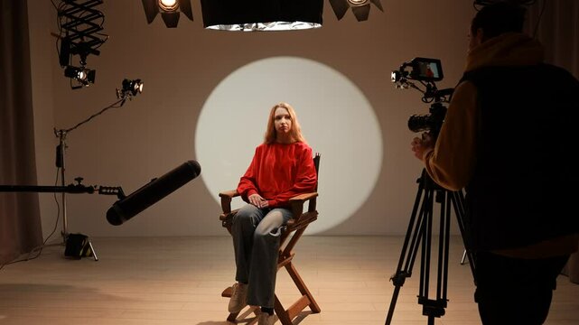Studio for photo video production filming with lighting equipment and cyclorama, model actress sits in front of camera on chair and talking, recording interview.