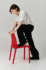 A handsome man in casual attire poses creatively on a red chair in a minimalist setting.