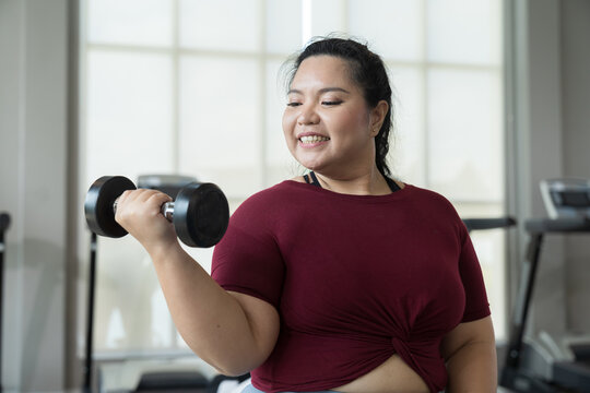 Happy smiling Asian young plus size woman lifting dumbbell weight in fitness club. Young overweight woman holding dumbbell workout at gym. Asian woman exercise training with weight, healthy lifestyle