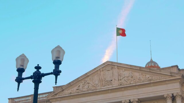 Street lamp in Lisbon. The Portuguese flag flutters in the wind atop the roof of a historic building, standing out against a vivid blue sky with a distinct plane trail in the background. 