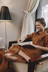 Relaxed woman in a cozy brown dress reading a book by a lamp in a modern living room, exuding...