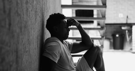 Man of African descent sitting against wall, arms wrapped around knees, eyes closed, deep in thought, exhaustion, mental burden, emotional distress, personal reflection