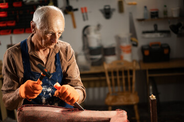 Furniture restoration concept. Elderly man removing old upholstery from vintage chair working in his workshop