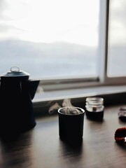 A cozy winter scene with steaming tea, a lit candle, and a knitted scarf resting on a wooden table by a window, creating a warm and inviting atmosphere