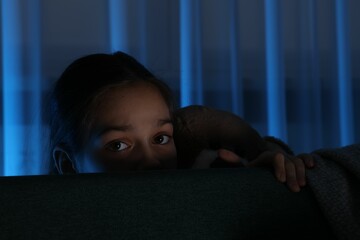 Scared girl with teddy bear hiding behind sofa at night