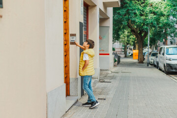 Young boy wearing a yellow vest and jeans stands on a city sidewalk, reaching up to press a...