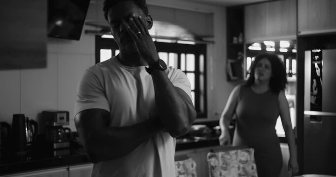 Husband standing in kitchen, hand on neck, looking away, wife in background talking, marital conflict, emotional distance, relationship tension, African descent