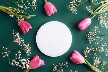 White round pedestal surrounded by pink tulips and baby's breath flowers on a green background. Top view, flat lay, mockup