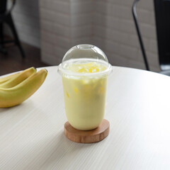 A light yellow smoothie is placed on a wooden coaster next to fresh bananas in a cozy cafe with modern decor