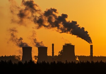 Smoking power plant silhouetted against the golden sunrise with dark trees below
