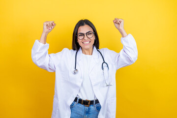Young brunette doctor woman wearing stethoscope standing over isolated yellow background very happy and excited making winner gesture with raised arms, smiling and screaming for success.