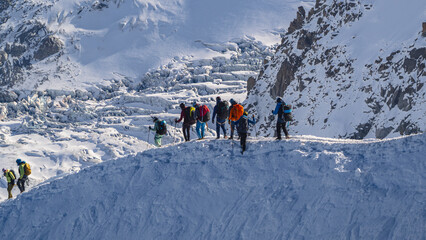Paragliders with large backpacks with a parachute inside descend a steep snowy slope from the summit of the Aiguille du Midi to make a parachute jump. 