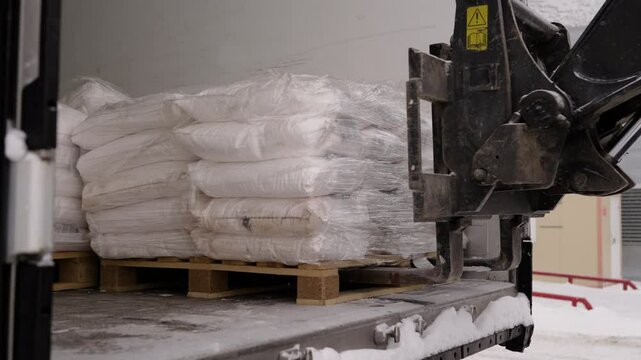 Winter logistics: forklift loading white sacks onto truck pallet at snowy warehouse loading dock during cold working day