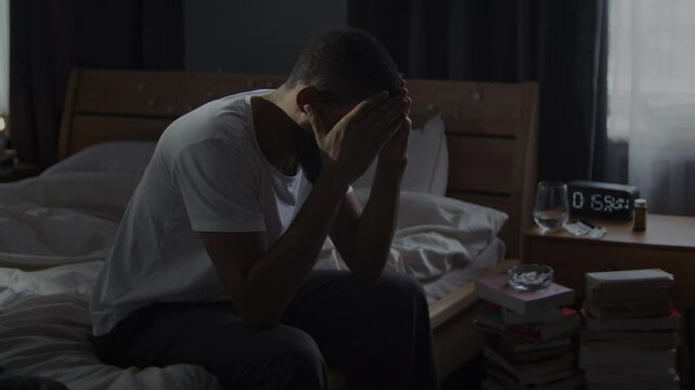 Black bearded man cries hiding face in palms on messy bed