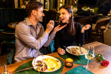 Young, beautiful couple feeds each other with food in a restaurant on a romantic dinner date.