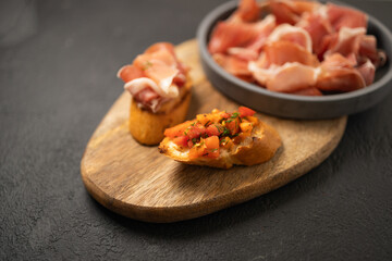 Sandwiches bruschetta with olives, bran bread, cherry tomatoes, prosciutto on wooden cutting board, dark black background surface