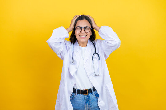 Young brunette doctor woman wearing stethoscope standing over isolated yellow background suffering from headache desperate and stressed because pain and migraine with her hands on head