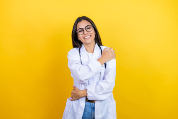 Young brunette doctor woman wearing stethoscope standing over isolated yellow background hugging...