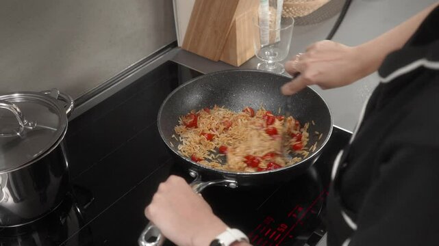 A woman cooks orzo pasta with shrimp and cherry tomatoes on an induction hob. The girl mixes the ingredients of the dish with a spatula in a non-stick frying pan. A housewife prepares a seafood dish.