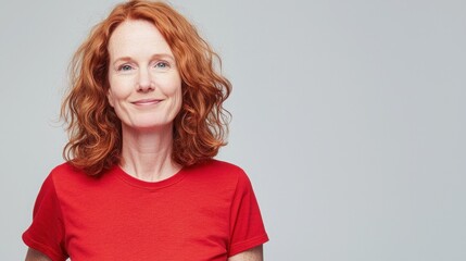 A middle-aged woman with vibrant red hair smiles gently against a light gray background, wearing a simple red t-shirt.