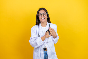 Young brunette doctor woman wearing stethoscope standing over isolated yellow background smiling with her hands on her chest and grateful gesture on her face.