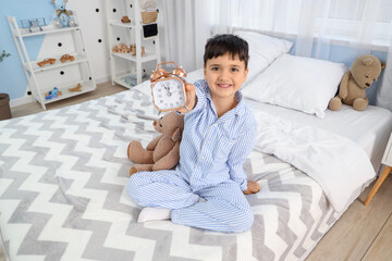 Cute happy little boy with teddy bear and alarm clock sitting in bedroom