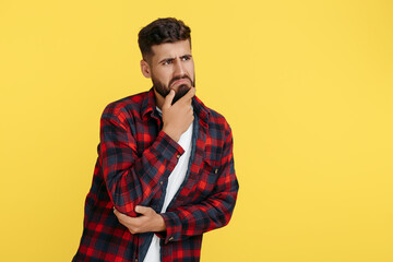 Confused bearded hipster young man in plaid shirt thinking over yellow background