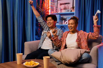 Couple gamer raising fist up holding joysticks be winner together complete level sitting sofa playing video game with front snack and drinks at neon light color living room at modern home. Infobahn.
