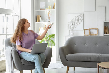 Young woman with air conditioner remote control and laptop sitting on armchair in living room