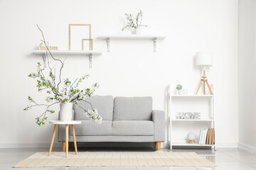 Vase with blooming branches on coffee table near soft sofa in white living room