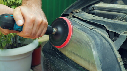 A close-up a person using a power drill with a buffing attachment to restore a cloudy and discolored car headlight. The headlight lens is being polished to improve its clarity and appearance.