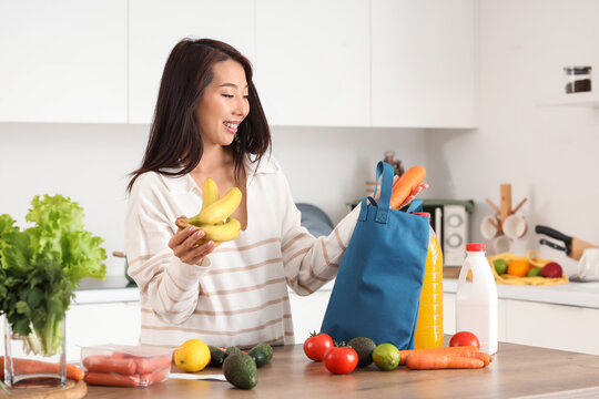 Young Asian woman unpacking groceries from blue shopper bag onto counter in kitchen