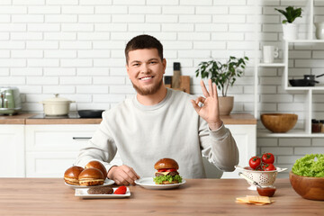 Young man with tasty burgers showing OK in kitchen