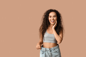 Surprised young African-American woman in loose jeans on beige background. Weight loss concept