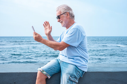 Cheerful senior man with beard sitting on sea shore using cellphone for video call, elderly pensioner enjoying freedom, vacation or retirement. Horizon over water